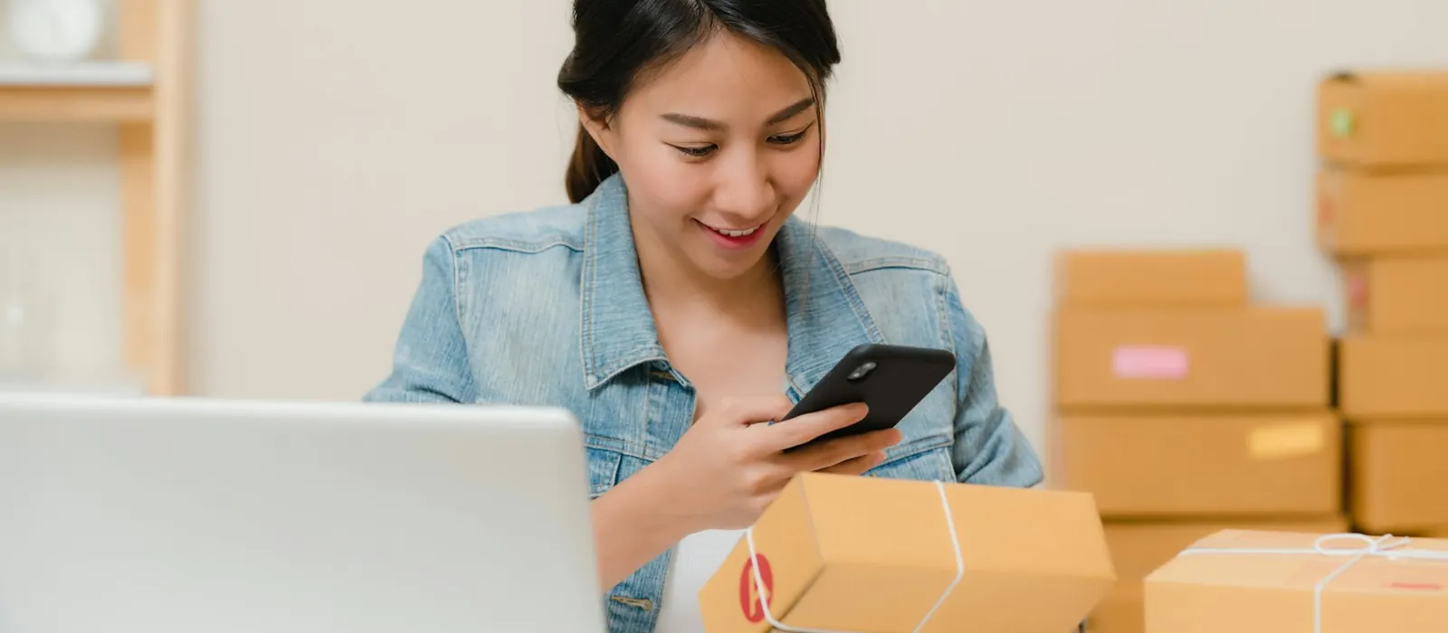 Woman checking a parcel with her phone beside a laptop and boxes