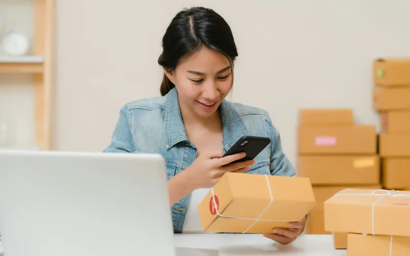 Woman checking a parcel with her phone beside a laptop and boxes